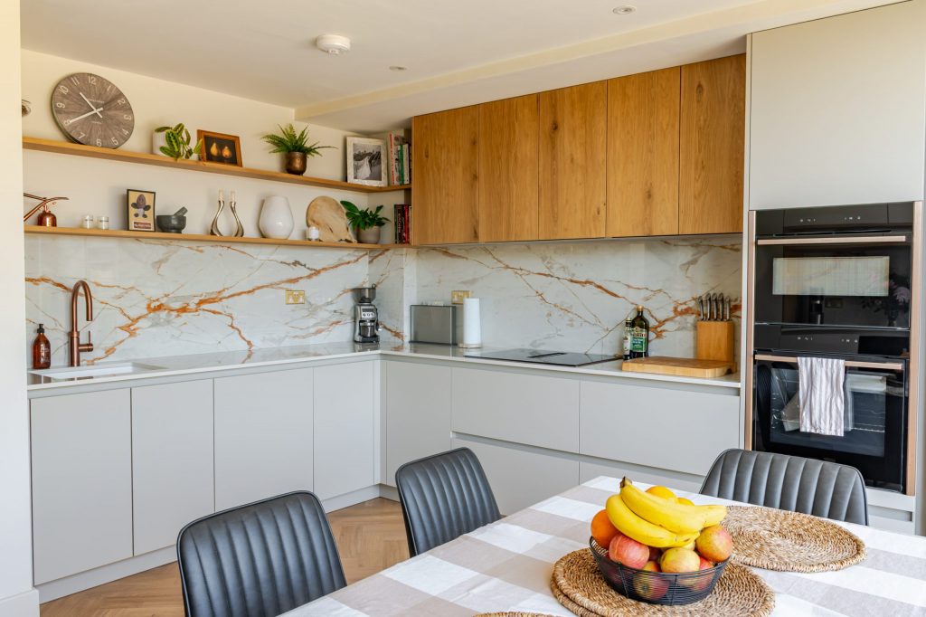 Wide angle showing floating shelves, Caple sink, Neff ovens, dining table, worktop and splashback in a cashmere and oak veneer Rotpunkt kitchen, Victoria Kitchens