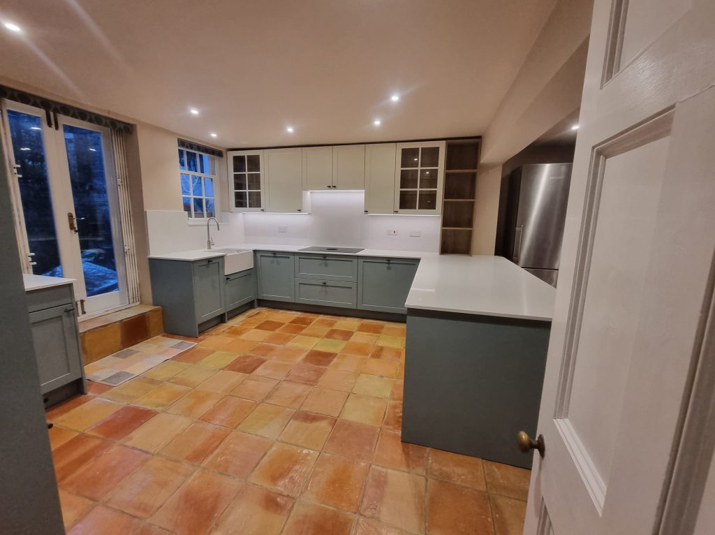 Full kitchen view showing two-tone cabinetry, oak shelving corner, integrated appliances and sink under window, Victoria Kitchens